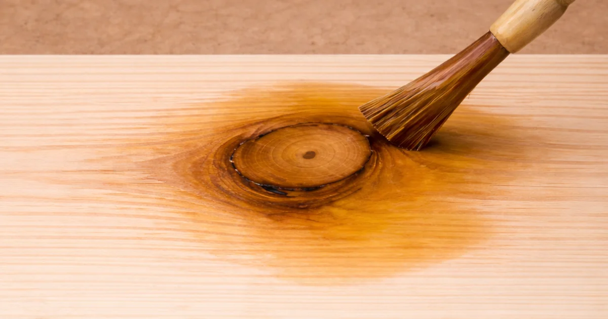 A brush applying clear treatment over a knot in a bare wood board.