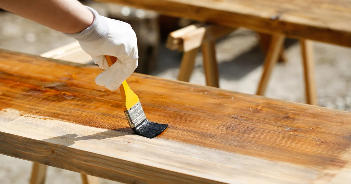 A gloved hand brushing a sealing coat onto a bare wood board outdoors.