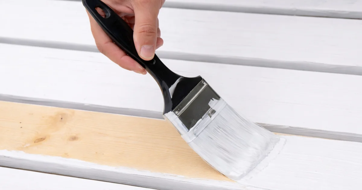 A hand using a wide brush to apply white paint to unfinished wood boards.