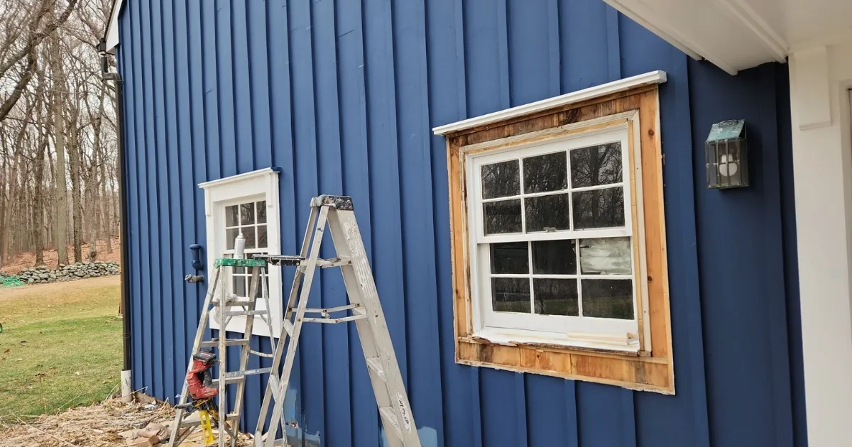 Bare wood window trim exposed for repair beside blue vertical siding.