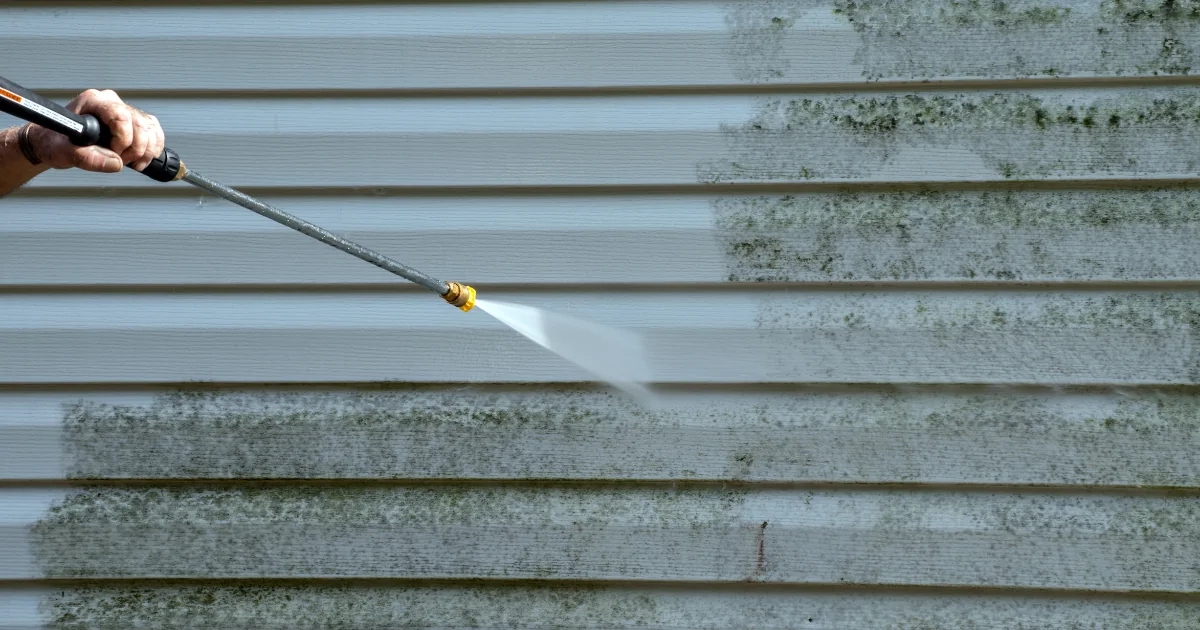 A pressure-washer wand removes dark mildew from light-colored horizontal siding.