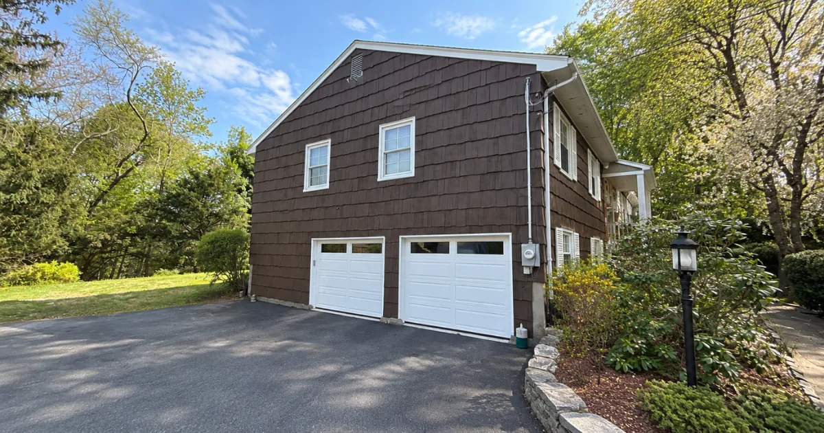 The same colonial after exterior work with a rich brown finish and white garage doors.
