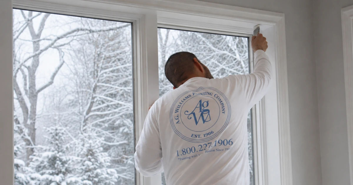 A.G. Williams painter in a branded white shirt finishing interior window trim with snow-covered trees outside